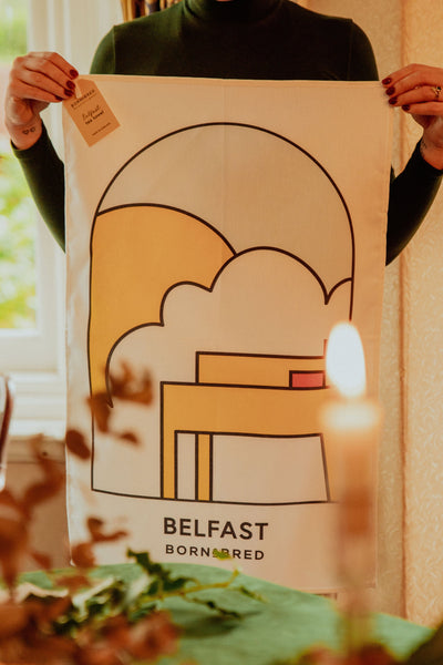 Person holding a poster with abstract design and 'Belfast' text, surrounded by natural light.