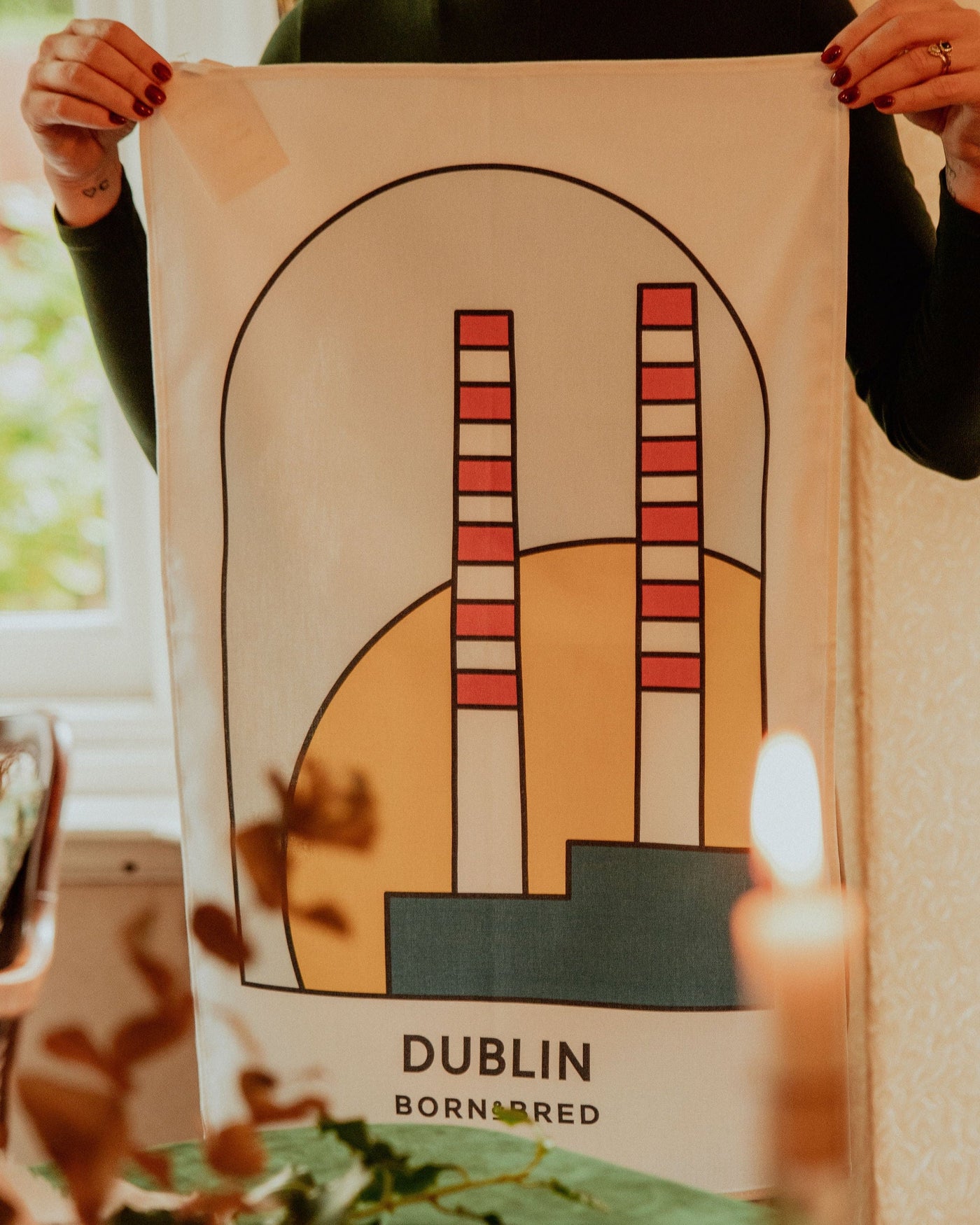 Person holding a poster with abstract design and 'Dublin' text, surrounded by plants and a candle.