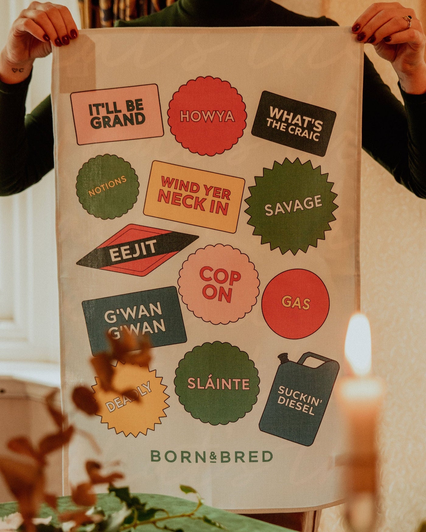 Person holding a decorative towel with colorful text and brand name 'Born & Bred' on a table.