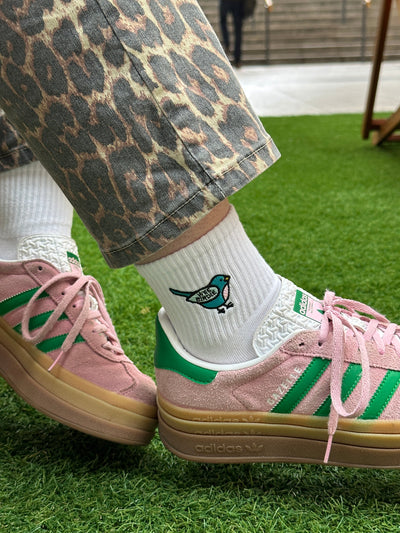 A person wearing patterned pants and white 'Wee Birdie' ankle socks with a bird design, standing on grass beside a pair of pink sneakers.
